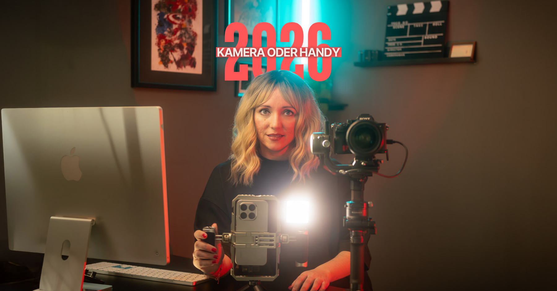 Blog-Banner mit dem Text „2026 KAMERA ODER HANDY“. Eine Frau sitzt an einem Schreibtisch vor einem Apple-Monitor. Vor ihr stehen eine professionelle Sony-Kamera auf einem Gimbal und ein Smartphone in einem Video-Rig mit eingeschalteter Leuchte.